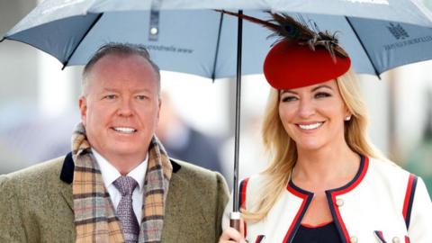 A photograph of Doug Barrowman and Baroness Michelle Mone at the Cheltenham horse racing festival in 2019. She is carrying an umbrella and wearing a red hat and white suit. He is wearing a light grey coat and tweed scarf. 