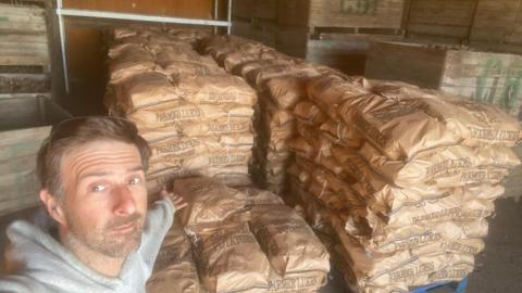 Luke a man with short brown hair, standing in front stacks of brown bags filled with potatoes. 