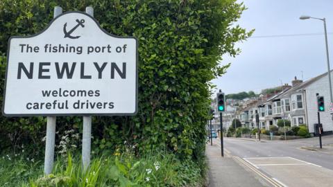 The welcome sign of Newlyn. It says the fishing port of Newlyn welcomes careful drivers. A green bush is behind the sign. To the right of the sign is a view down a street in the town. A row of houses and traffic lights are visible. 