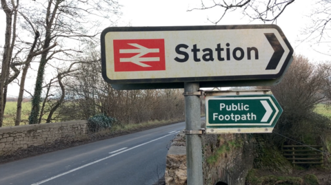 A road sign for a train station above a green public footpath sign on a pole by a roadside in Devon. It is next to a stone bridge in the countryside.