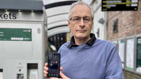 Justin Baker with short white hair and glasses, holding a phone to the camera and wearing a blue sweater and black shirt. He is standing by a railway ticket machine. A departure screen is just visible in the upper right of the picture. Departure boards can also be seen on a brick station wall.