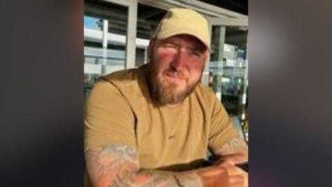 A man wearing a tan coloured baseball cap and t-shirt is sat looking into the camera. He has tattoos on both arms and a brown beard.