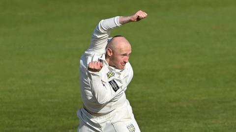 Left-arm spin bowler Callum Parkinson just after completing a delivery with his left arm tucked in and his right arm raised over his head