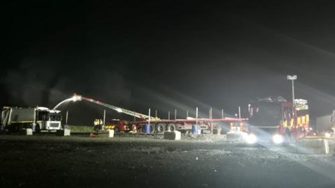 The long crane from a fire service appliance is hosing water over a fire. There is some smoke in the background and a fire engine in the foreground with its lights on as it is dark.