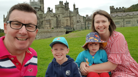 Darren Wheatley mewn crys streipiog pinc, Oscar, 7 mewn het a siwmper glas, Ffion, 2, mewn het batrymog a chardigan glas. Nicola Wheatley yn dal Ffion mewn ffrog binc a Gwyn. Y pedwar yn gwenu o flaen castell.