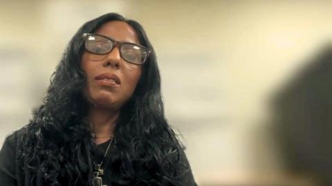 Shira Javaid has a serious expression on her face as she looks towards the camera. She has long, wavy black hair and wears a black top, large, dark-rimmed glasses and a silver necklace with pendants.