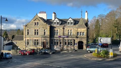 A general view of the NatWest bank in Windermere. The three-storey building is Victorian in style with a typical Cumbrian slate roof. There are cars parked outside it and the sign for Windermere town centre is visible at the nearby junction.