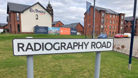 A street sign which reads "radiography road" in front of red brick buildings.