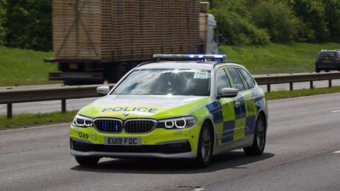 An Essex Police car is parked in the middle lane of a three-lane carriageway. It is the only vehicle on that side. On the opposite side there is a lorry and car driving past.