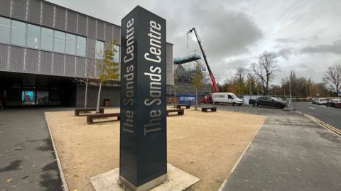 A general view of the entrance to The Sands Centre in Carlisle. A black pillar has the name of the venue written on each side in silver. The entrance to the leisure centre is visible on the left, while on the right heavy plant machinery is working on an area of the building surrounded by scaffolding.