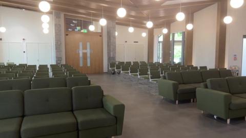 A photo shows eight rows of chairs on the left hand side of the picture, and a walkway in the middle, and eight rows on the other side of chairs. The chairs are green. There is also double doors in the middle which are closed, and round lights hanging from the ceiling omitting a pale white light. 