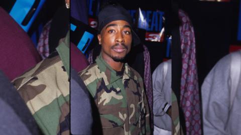 Image of Tupac Shakur wearing a blue bandana and a camouflage jacket.