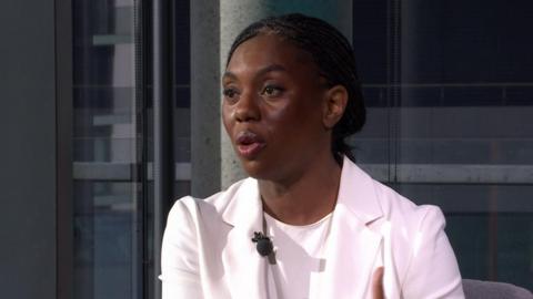 Kemi Badenoch in White blazer and shirt sitting down on a chair