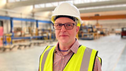 A man stands on a factory floor wearing a hardhat and glasses, as well as a hi-vis jacket. He also wears glasses.