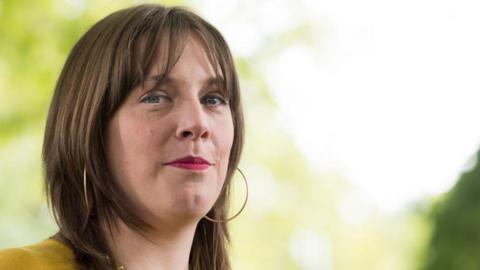 Jess Phillips has short brown hair. She is wearing a yellow top and is stood looking to the right of the image.