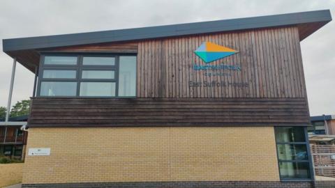 Outside of East Suffolk Council building. It has a sloping roof and wood panelling to the first floor and the outside of the ground floor is light-coloured brickwork. The council logo is on the wooden section