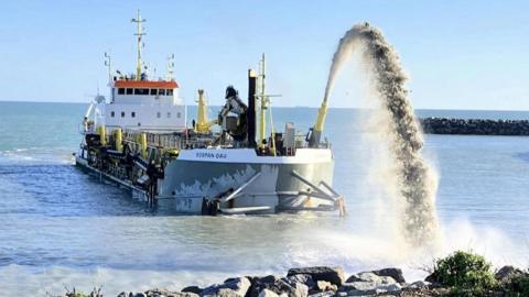 A boat in a body of water firing shingle into an arch which is landing on a coastline.