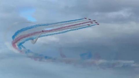 The Red Arrows, a formation of seven red planes in the sky with blue, white and red smoke trailing in an arching shape behind.