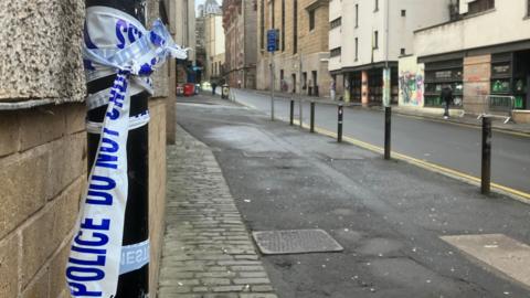 Broken police tape attached to a lamppost in Edinburgh's Cowgate. There are no vehicles on the road. The pavement is empty. There are several bollards between the pavement and road.