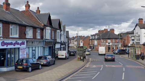 A general view of Derby Road in Stapleford, Nottinghamshire. Cars are parked at the side of the road next to a row of shops