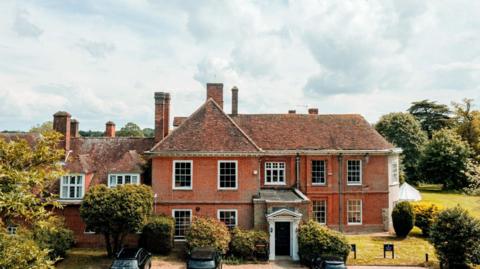 The main Stoke College building, made of red bricks with multiple chimneys. There are three vehicles parked in front of the building. Trees surround the building. 