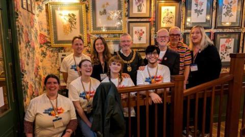 A group of Thame Pride organisers posing for a picture in front of a wall with flowery wall paper and colourful paintings