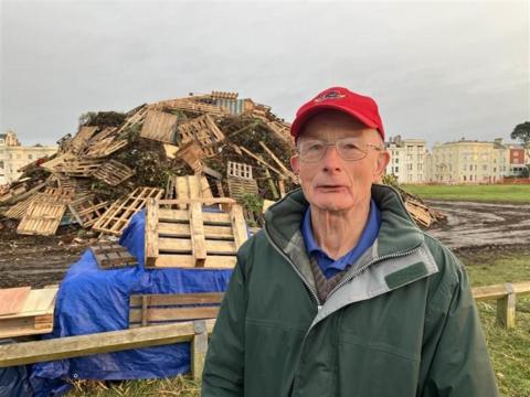 An older man in a green cagoule and red hay stands in front of a bonfire pyre which is being built. 