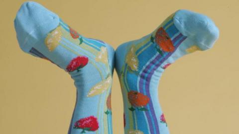 a pair of feet in the air with blue socks with colourful fruit pictured on them on a yellow background