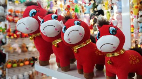 Four red plush horse toys are lines on a shelf in ascending height order. They have big blue eyes which are looking down and to the right. They each have a white muzzle with an upside down smiley face on it. It makes it look like the horse is sad. 