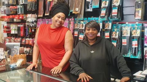 Two Women smiling at the camera, behind a shop counter with rows of hair products in the background.