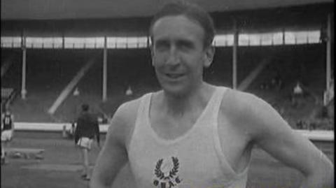 A black and white image of Alan close up to camera in a white athletic vest standing in a stadium with a couple of other athletes in the background and empty stands