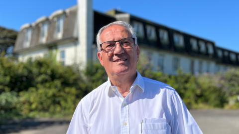 A man with grey hair and black glasses. He's wearing a white shirt standing in front of hotel, the St Margaret's Lodge. 