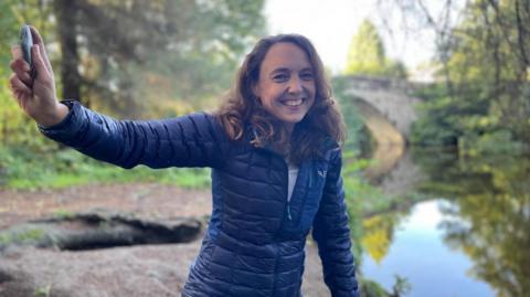 A woman with shoulder length brown hair smiles as she holds up a stone above her head. She's standing next to a river bank