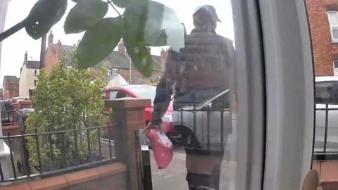 Doorbell camera footage of a man dressed in black with a black cap walking out of a front garden with a pink parcel.