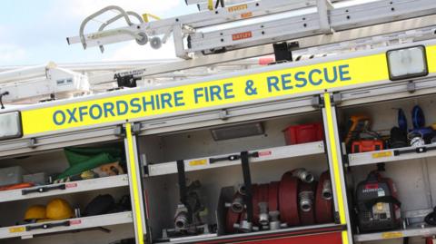 Viewed from the side, a fire engine has aluminium ladders stored on its roof. Oxfordshire Fire & Rescue is printed on the side in blue lettering on a birght yellow background. The shutters on the side of the vehicle are open, displaying rolled red hoses, yellow helmets and various tools stowed in compartments.