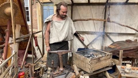 A man stokes a fire as he demonstrates how a Roman blacksmith would have worked.  He wears an old apron and a toga-style top tied at the shoulder.