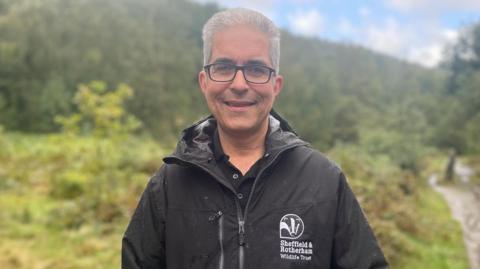 A grey haired man with dark glasses stands in a black waterproof jacket surrounded by bushes and trees