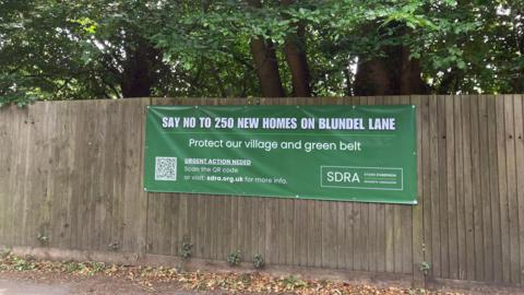 A green banner hanging on a fence which has the words "say no to 250 new homes on Blundel Lane" and a website and QR code on it.