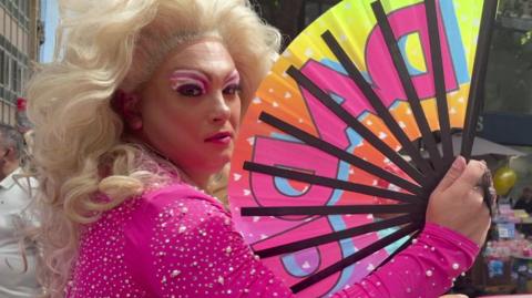 A drag queen walking through the centre of Norwich as part of the city's Pride March celebrations. They are looking directly at the camera and are wearing a pink pearl embellished outfit. They have pink makeup and are wearing a curly blonde wig. In their right hand they are holding a fan which is open and says DADDY on it.