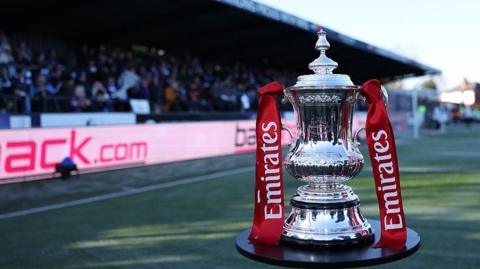 The FA Cup trophy pictured on a stand at Macclesfield's Moss Rose ground. There is a stand full of supporters in the background of the image. 