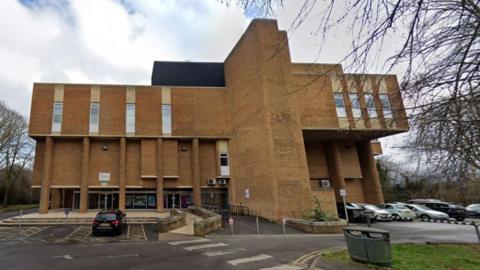 Peterborough's Regional Pool - a brutalist structure in an abstract shape, with small windows across the top level. In front of it are a number of parked cars.