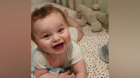 Baby Preston Davey smiles at the camera dressed in a white vest. He appears to be lying in a cot with a star-covered bedsheet.