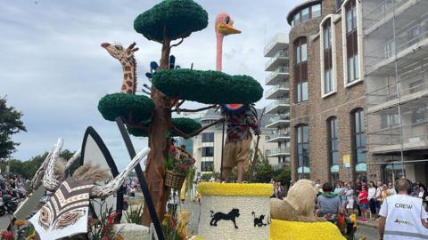 A picture of a float at the Battle of Flowers in 2025. There is a animal themed float with giraffes and lions. Spectators are lining the road.
