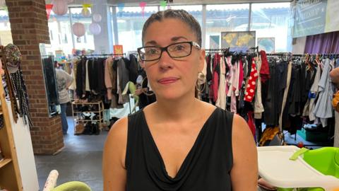 Tingy Simoes looks at the camera. She is stood in a large room with rows of clothes on racks.