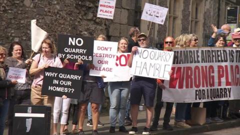 A group of protesters are lined up along the side of a road holding up signs saying 'no quarry' and 'don't destroy our village'. There are about 15 people in the image but there are more out of shot.