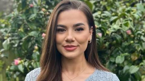 A woman with long brown hair, wore in a side parting. She is stood outside in a garden and wearing a blue knitted top.