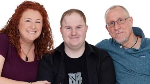 Catherine, Daniel and Jonathan in a family portrait. Catherine has red hair and is smiling. Daniel also has red hair and wears a black hoodie. Jonathan has short white hair and wears a blue polo shirt.