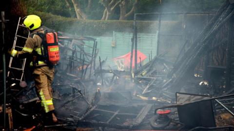 A firefighter stood in a badly fire-damaged garden. The photo shows the ruins of what could be a shed, as well as a charred wheelbarrow and a fire-damaged children's play house.