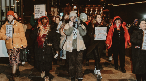 Women marching through Cardiff City Centre at night with placards and speakers, calling for and end t oall violence against women and girls