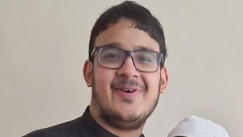 A young man with black hair, square black glasses, a beard around the edge of his jaw and a moustache. He is smiling and looks as if he might be midway through speaking. He is wearing a black shirt with a small collar.